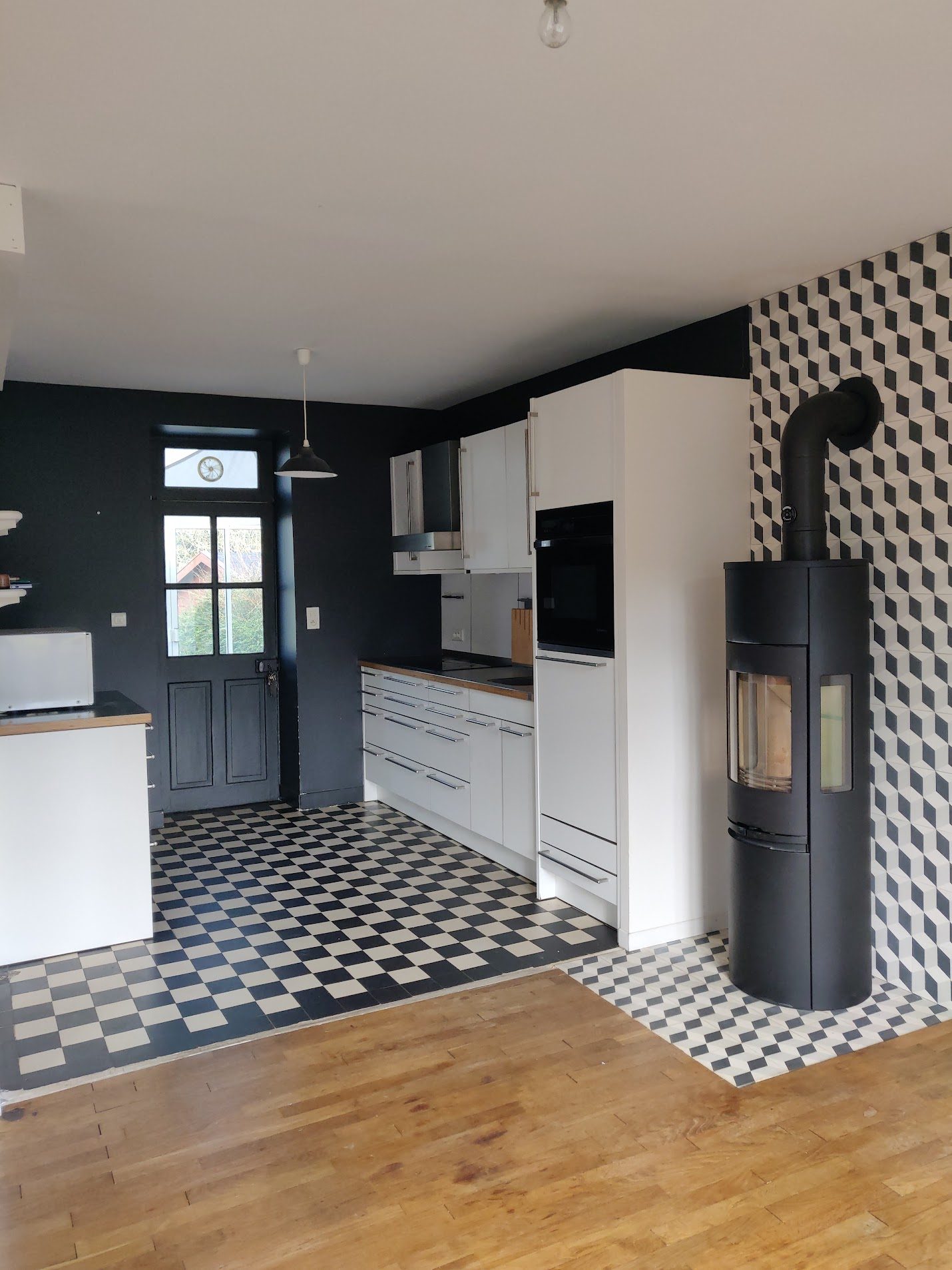 Cuisine avant rénovation : vue d'ensemble de l'ancienne cuisine avec murs ardoise, carrelage damier et poêle à bois