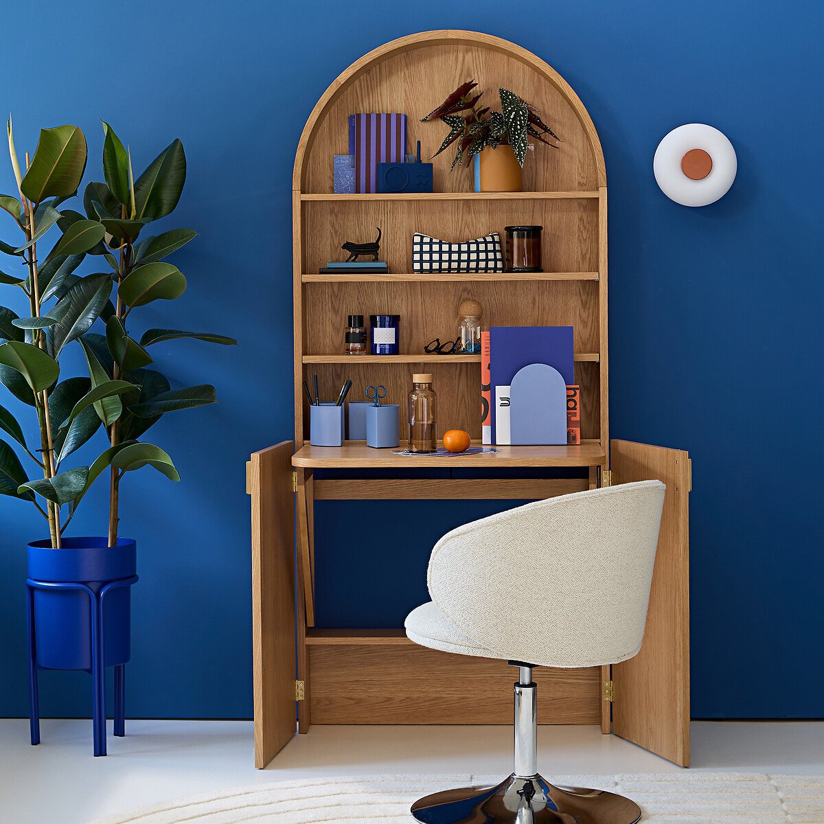 Bureau pliable Marcela en chêne, abattant ouvert contre un mur bleu avec un fauteuil en bouclette